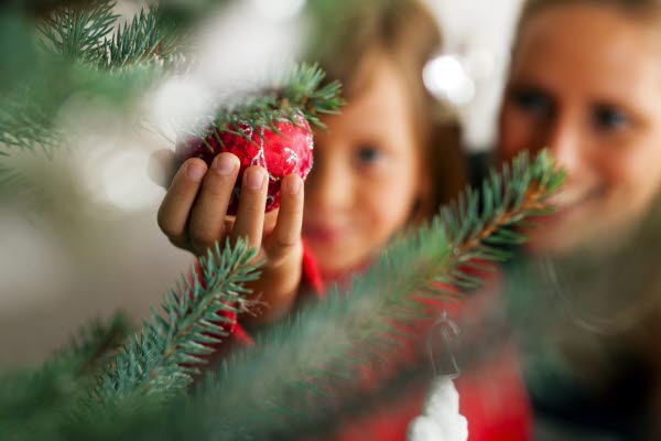 Ett barn håller i en röd julgranskula som hänger i en gran. Bredvid barnet sitter en kvinna.