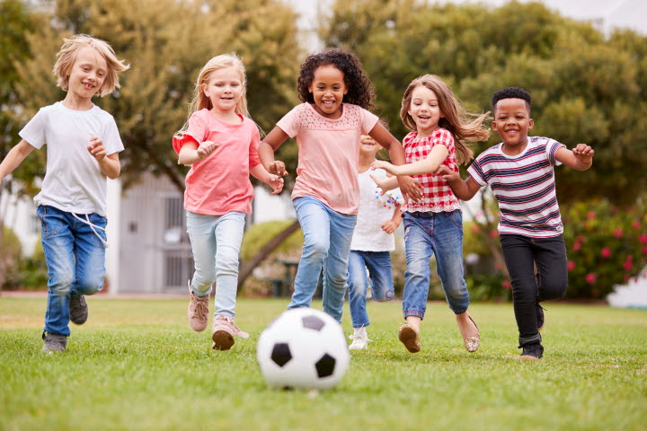 Flera glada barn springer på en grön gräsmatta mot en fotboll.