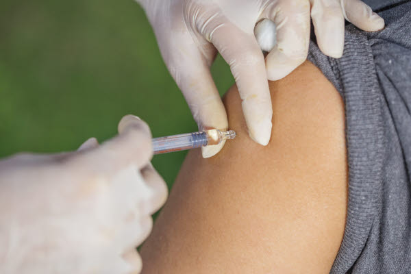 En person med grå uppkavlad tröjärm får en vaccinationsspruta i överarmen. En hand med vit plasthandske håller i sprutan. Bakgrunden är grön.