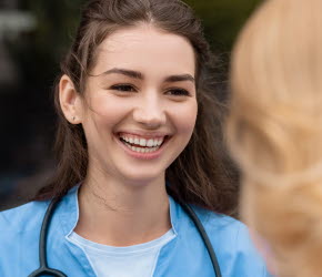 En kvinnlig sjuksköterska ler mot en rödhårig kvinna. Runt halsen bär sjuksköterskan ett stetoskop.