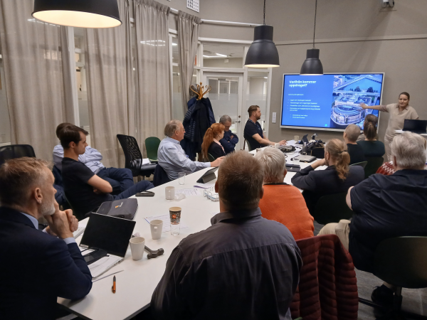 En grupp människor sitter vid ett konferensbord och tittar på en kvinna som håller föredrag.