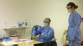 En kvinna sitter vid ett bord och instruerar en annan kvinna i hur antigentestning fungerar. Båda kvinnorna är klädda i blåa arbetskläder och har blåa gummihandskar på händerna. Över ansiktet har de ansiktsmask och visir.