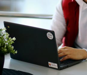 En elev i vit huvtröja och röd väst, sitter framför en svart Chromebook med händerna på tangenterna. 