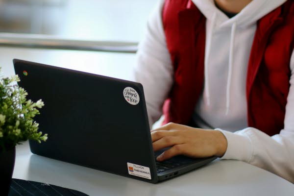 En elev i vit huvtröja och röd väst, sitter framför en svart Chromebook med händerna på tangenterna. 