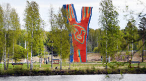 En bild över skulpturen Y:et vid Midlandaområdet. Runt omkring syns gröna träd, och i framgrunden skymtar man vatten.
