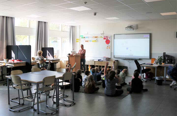 Klassrummet har bänkar som sitter ihop två och två längst med väggarna, i mitten är det ett gruppbord och på govlet sitter barnen på en matta under genomgången.