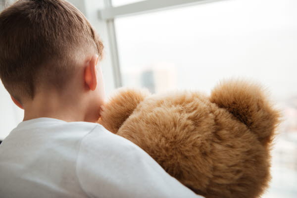 En liten pojke sitter med ryggen emot och håller i en brun nallebjörn. Pojken har en vit långärmad tröja och tittar ut genom ett fönster.