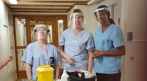  Syntolkning av bild: Tre kvinnor iklädda blåa arbetsskjortor och med visir över ansiktena. Framför står en vagn med olika redskap och tillbehör för att kunna utföra vaccinering.