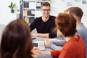 En man i glasögon ler mot en grupp kollegor som sitter vid ett skrivbord.
