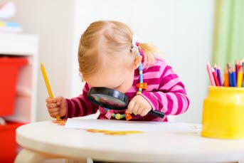 Flicka som sitter vid ett lekbord och tittar genom ett förstoringsglas samtidigt som hon håller i en penna.