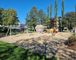 En lekplats med sandlåda, gungor och en klätterkub med rutschkanor. Lekparken är omgiven av höga träd, solen skiner och himlen är blå.
