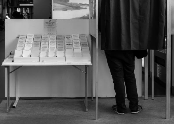 En person står i ett bås med ett skynke fördraget och röstar. Till vänster om båset står ett bord fyllt med valsedlar.