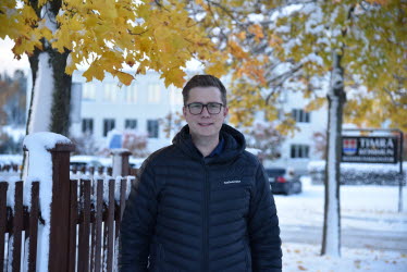 En man står utanför Timrå kommunhus. Snön ligger vit runtomkring och trädets löv har starka höstfärger i färgskalan gult. I bakgrunden skymtas en skylt där det står "Timrå kommun".