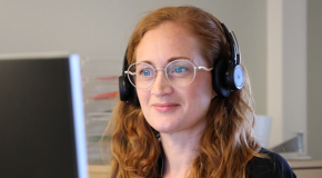 En kvinna med rött lockigt hår och glasögon sitter och arbetar vid en datorskärm. På huvudet har hon svarta hörlurar.