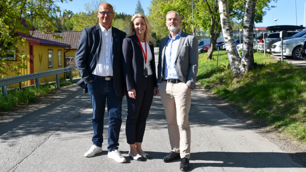 g	Tre personer står uppradade på en väg och tittar in i kameran.