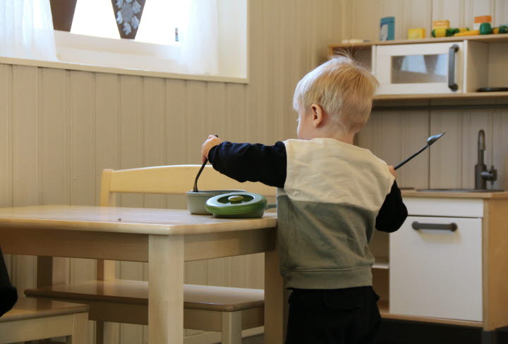 En pojke runt 1 år står vid ett barnbord och leker med en spade, slev och kastrull. I bakgrunden står ett minikök anpassat för barn.