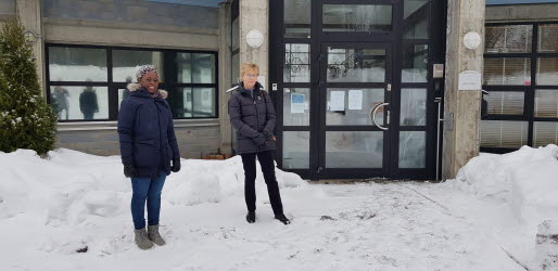 Två kvinnor står framför en byggnad med fönster och en stor port med glas i bakgrunden. Kvinnorna har mörka vinterjackor på sig och marken är täckt av snö.