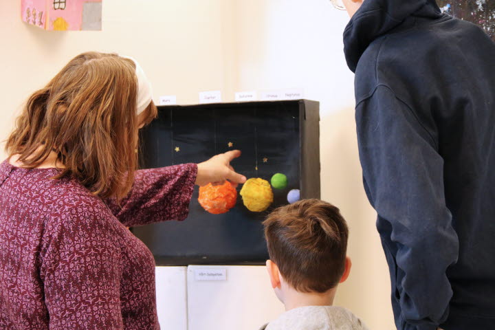 En vuxen och två barn tittar på olika konstverk som eleverna på grundsärskolan i Bergeforsen skapat. Den vuxna pekar på kostverket.