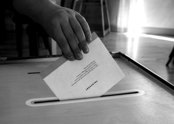 En svartvit bild där en hand lägger ner ett röstkort i en trälåda. På bilden ser man texten val 2022