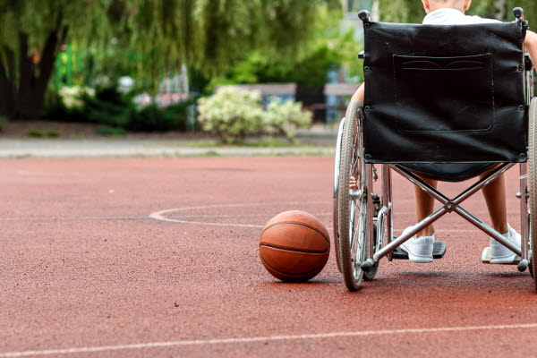 Ett barn sitter i en rullstol på en basketplan. Bredvid honom ligger en orange basketboll.