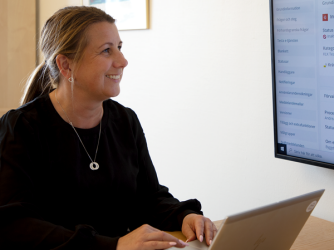 En kvinna i svart tröja och uppsatt hår sitter vid ett skrivbord med händerna på en laptop. Hon tittar glatt upp mot en skärm och ler.