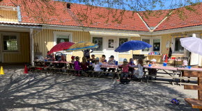 Barn och pedagoger sitter vid ett långbord på förskolans innergård. Barnen sitter under färgglada parasoll. Skuggor av grönskande träd mot marken. 