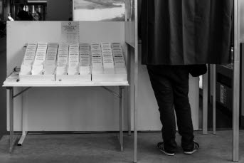 En person står i ett bås med ett skynke fördraget och röstar. Till vänster om båset står ett bord fyllt med valsedlar.