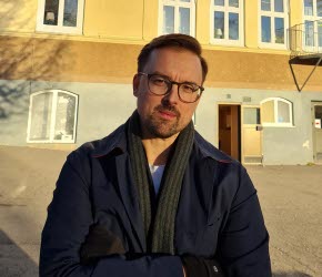 En man med glasögon står framför en gul skolbyggnad. På byggnadens fasad sitter en skylt texten "Fagerviks skola". 