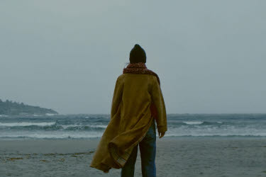 En kvinna i orange jacka står vänd mot havet