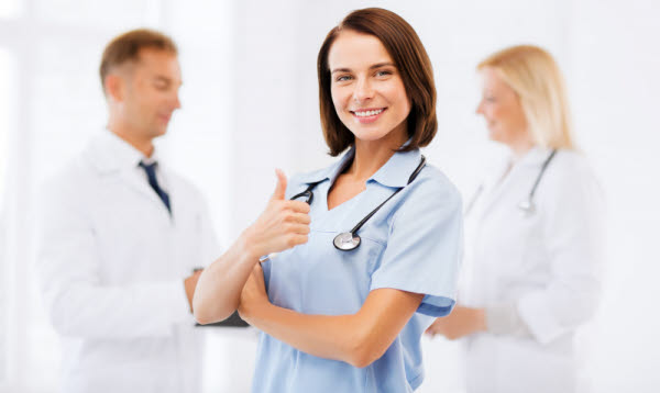 Bilden föreställer en sjuksköterska som gör tummen upp. I bakgrunden syns två medarbetare som pratar med varandra. 