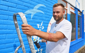 John står på en stege och håller i en graffitiburk vid sin väggmålning.