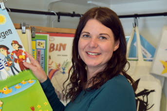 En kvinna håller upp en barnbok och tittar leendes in i kameran.