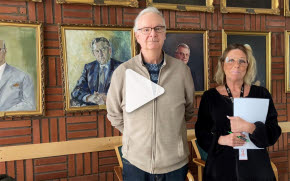 En man och en kvinna står framför porträtt av tidigare kommunfullmäktigeordföranden. Kvinnan håller dokument i sina händer.