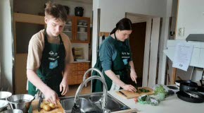 Två elever med förkläden står vid en köksbänk med varsin skärbräda. Den ena hackar potatis och den andra hackar örter.