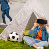 Två barn i ett lekrum. Ett av barnen sitter ner och har på sig VR-glasögon. Andra barnet åker skateboard i bakgrunden.