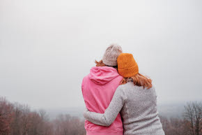 Två kvinnor håller om varandra. Kvinnan till vänster har rosa tröja och grå mössa. Kvinnan till höger har grå tröja och orange mössa.