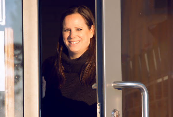 En kvinna står öppnar en dörr till en utomhusentré. Hon ler och har solen i ansiktet.