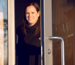 En kvinna står öppnar en dörr till en utomhusentré. Hon ler och har solen i ansiktet.