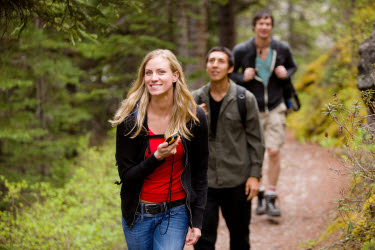 En kvinna med en GPS i handen promenerar i skogen. I sällskap bakom henne kommer två män gåendes. 
