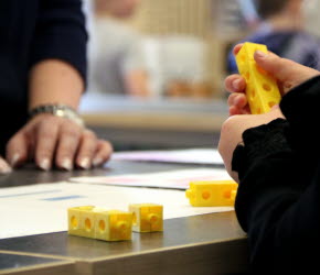 Ett barn håller i gula byggklossar och sitter vid ett bord i ett klassrum. På bordet ligger papper och fler gula byggklossar.