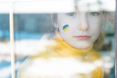 En flicka i gul stickad polotröja tittar in i kameran med ledsen blick. På kinden har hon en Ukrainsk flagga målad i blått och gult.