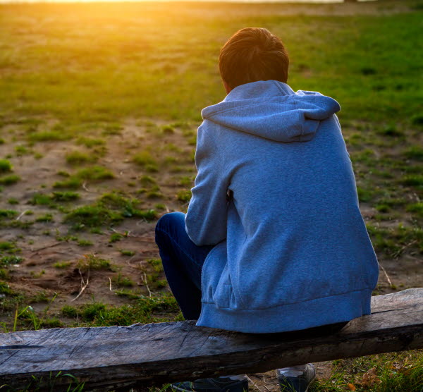 Tonåring i blå tröja sitter på en bänk och tittar mot solnedgången