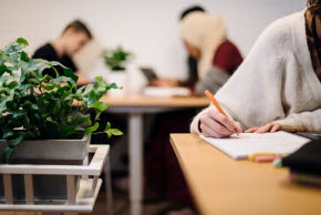 En student med vit kofta sitter vid ett skrivbord och skriver på ett papper. Bredvid henne står en grön växt i en kruka.