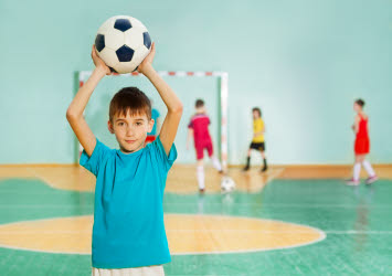 Pojke i blå tröja håller i fotboll med armarna ovanför huvudet. Barn i bakgrunden spelar fotboll.
