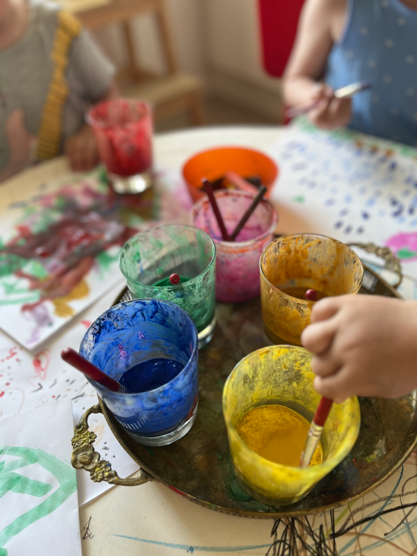 Flera glas med målarfärger står på en bricka. Ett barns hand rör med pensel i den gula färgen.