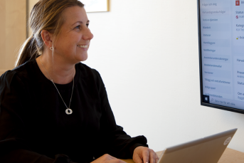 En kvinna i svart tröja och uppsatt hår sitter vid ett skrivbord med händerna på en laptop. Hon tittar glatt upp mot en skärm och ler.