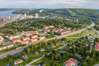 En vy av en stad omgiven av grönska och vatten, med byggnader, vägar och fjärran kullar. Utsikten visar både bostadsområden och industriella anläggningar vid vattnet.