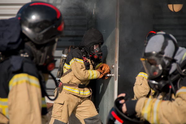 Tre elever i brandskyddsutrustning står framför en byggnad. Rök väller ut ur en dörr.