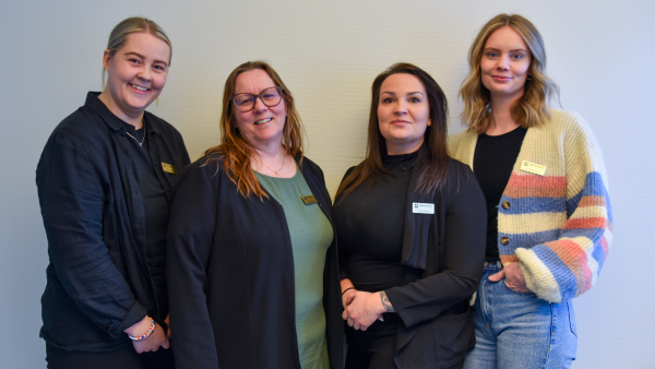 Fyra kvinnor står på rad och tittar in i kameran och ler.