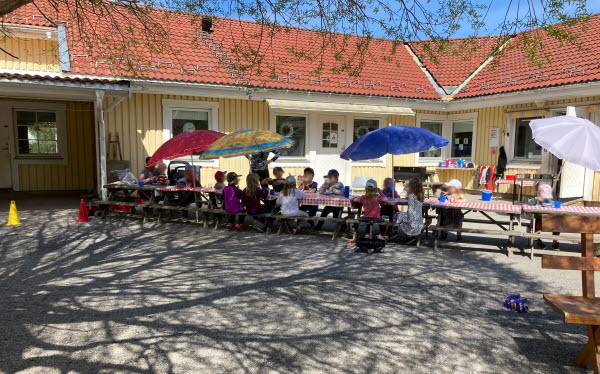 Barn och pedagoger sitter vid ett långbord på förskolans innergård. Barnen sitter under färgglada parasoll. Skuggor av grönskande träd mot marken. 